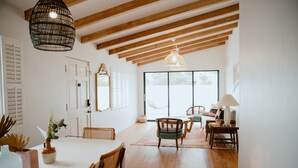 A spacious room with wooden beams, a dining table with chairs, a large window, and a hanging light fixture.