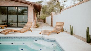 A pool area with two wooden lounge chairs, a brick building, and a white wall with cacti.