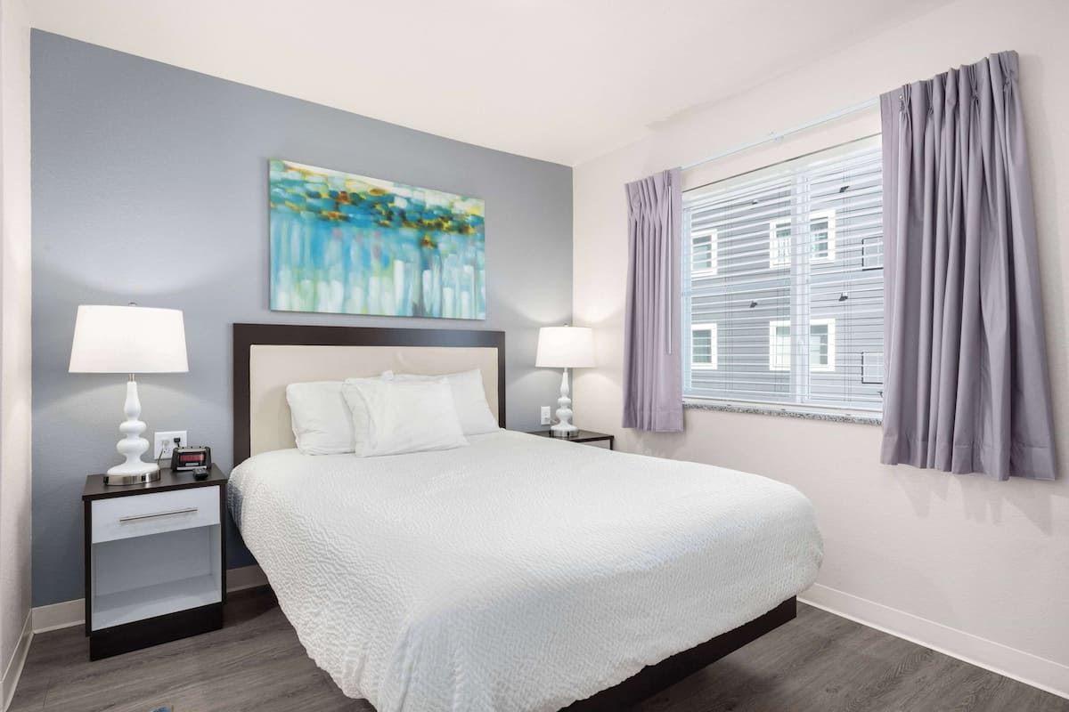 A neatly arranged hotel room with a large bed, two bedside tables with lamps, a painting on the wall, and a window with curtains.