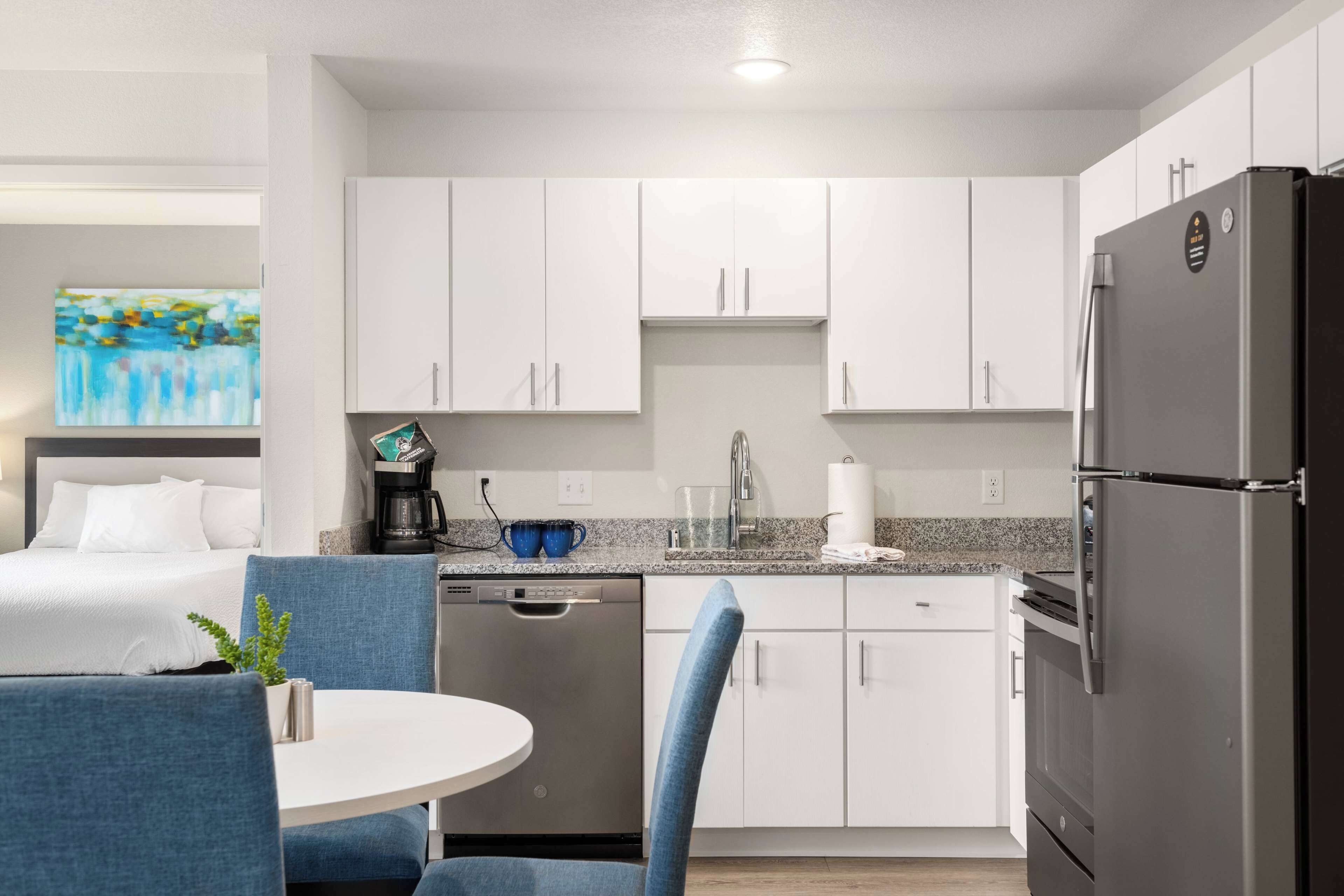A modern hotel room with a bed, a small dining area with a table and chairs, a kitchen with white cabinets, a refrigerator, and a coffee machine.