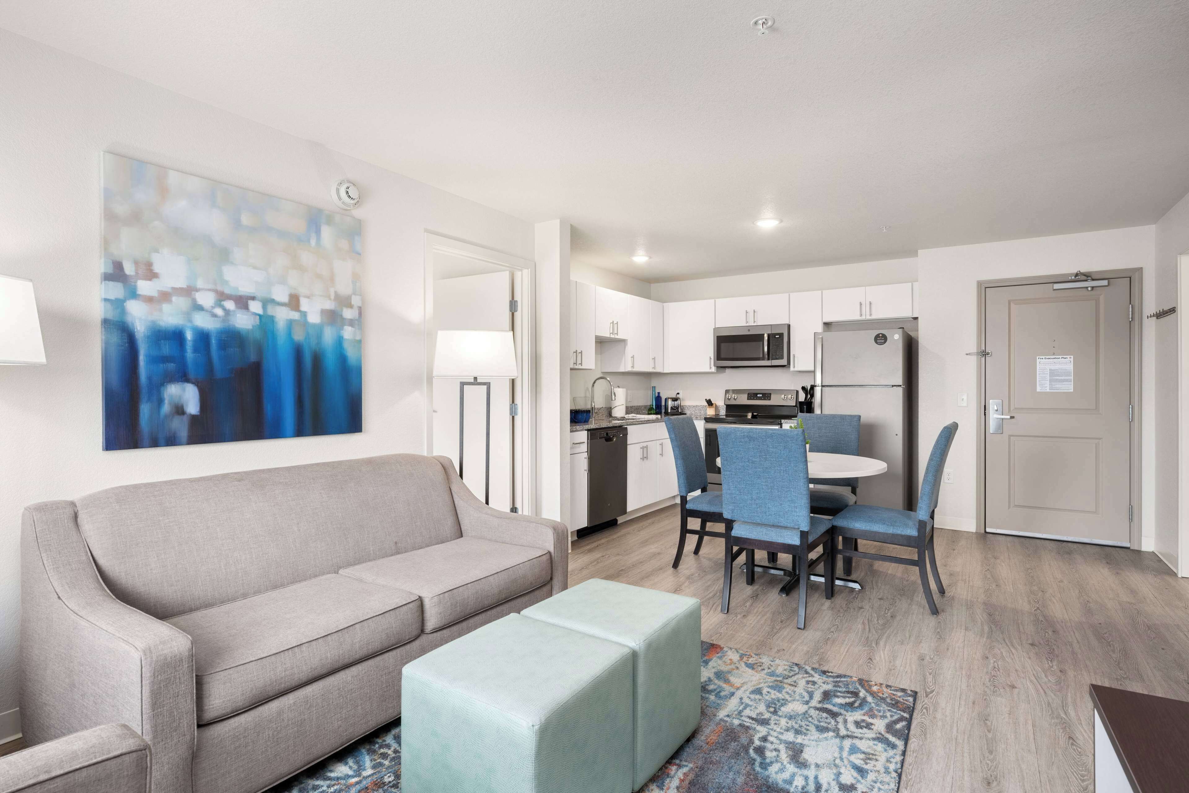 Een moderne woonkamer met een grijze bank, een klein voetenbankje en een eethoek met blauwe stoelen.