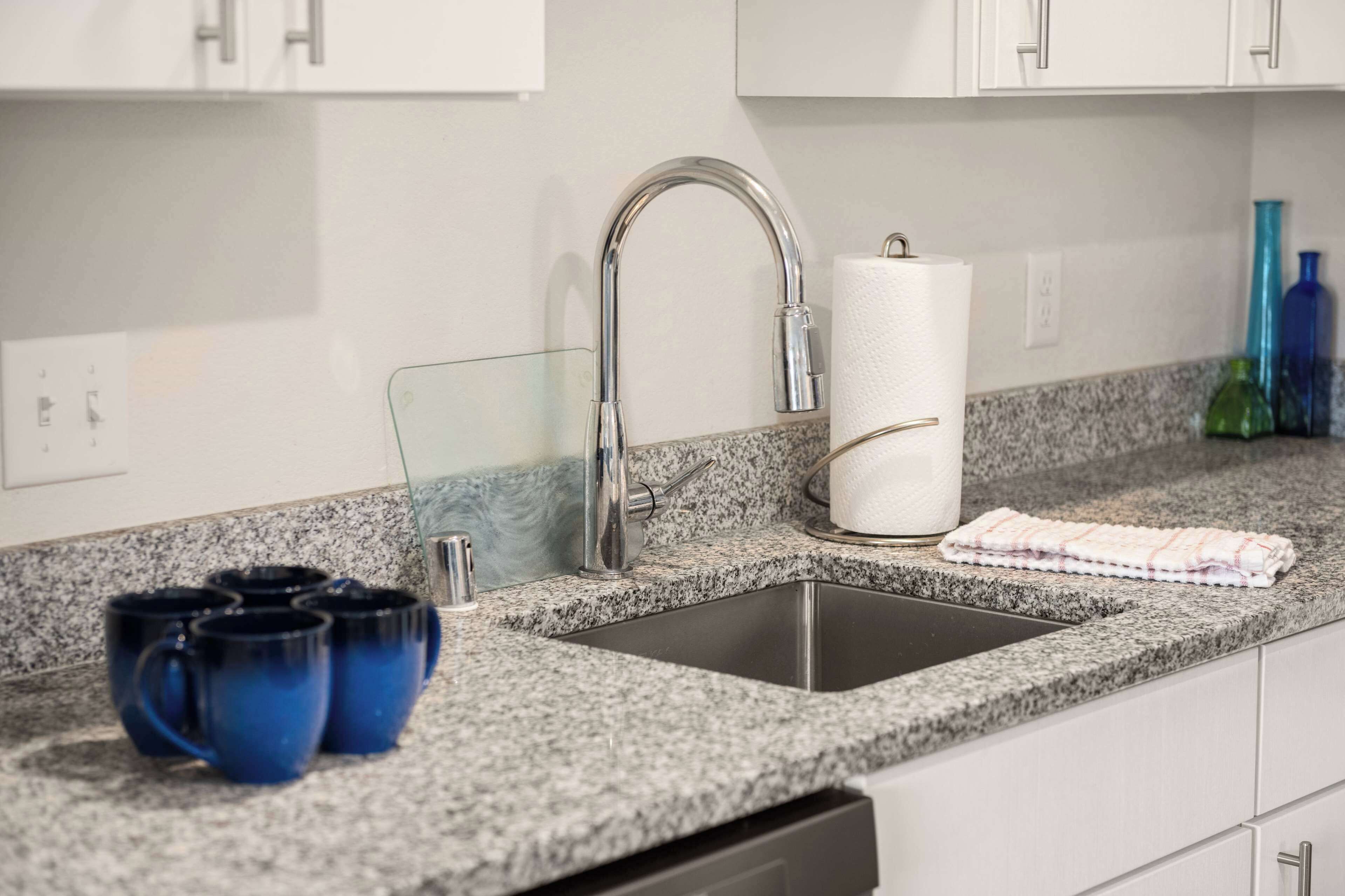 A kitchen with a granite countertop, stainless steel sink, and white cabinets.