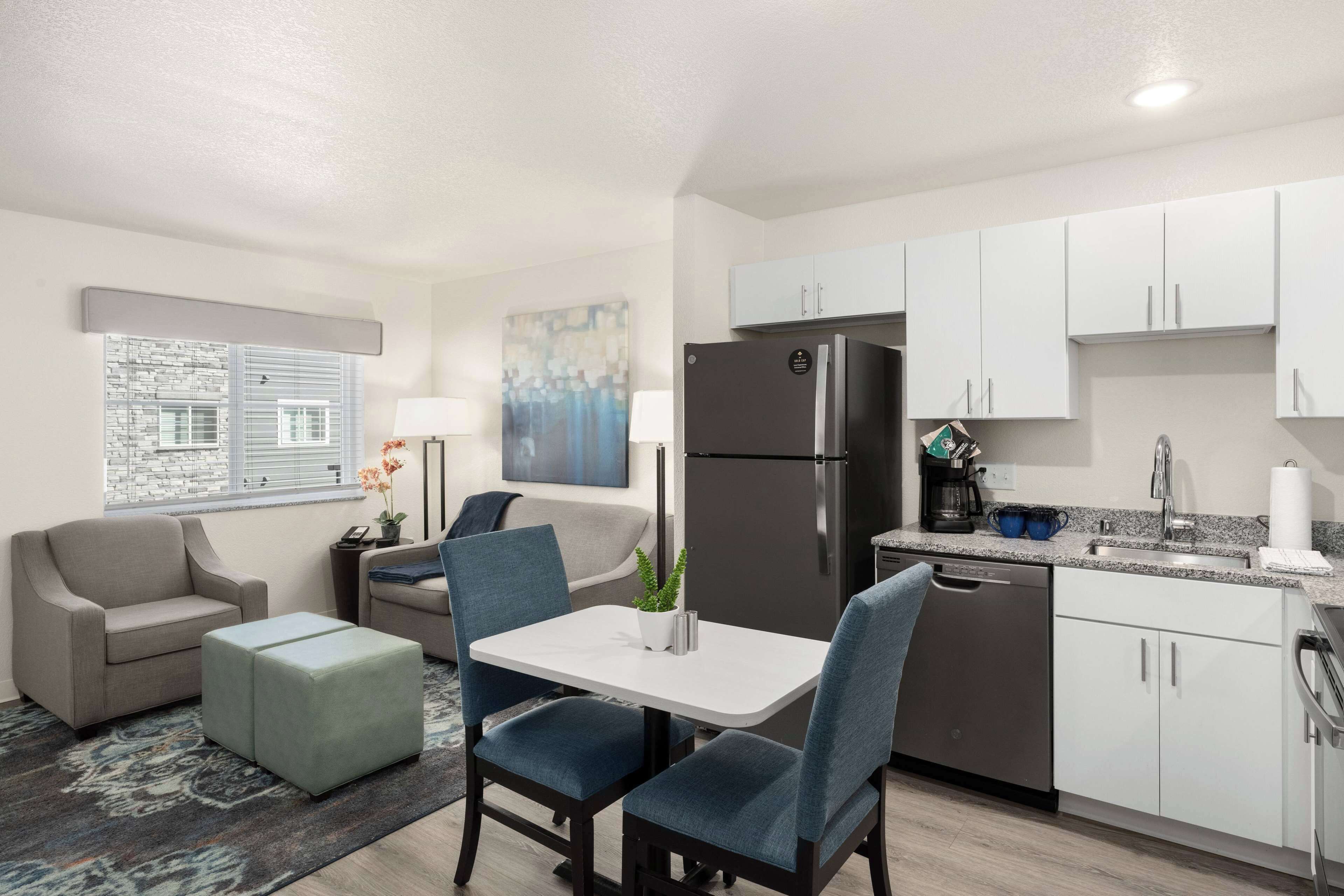A compact living space with a kitchenette, a dining area with a table and chairs, a sofa, a small coffee table, and a window with blinds.