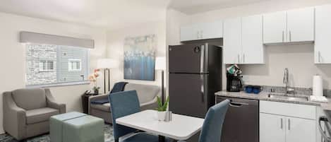 A compact living space with a kitchenette, a dining area with a table and chairs, a sofa, a small coffee table, and a window with blinds.
