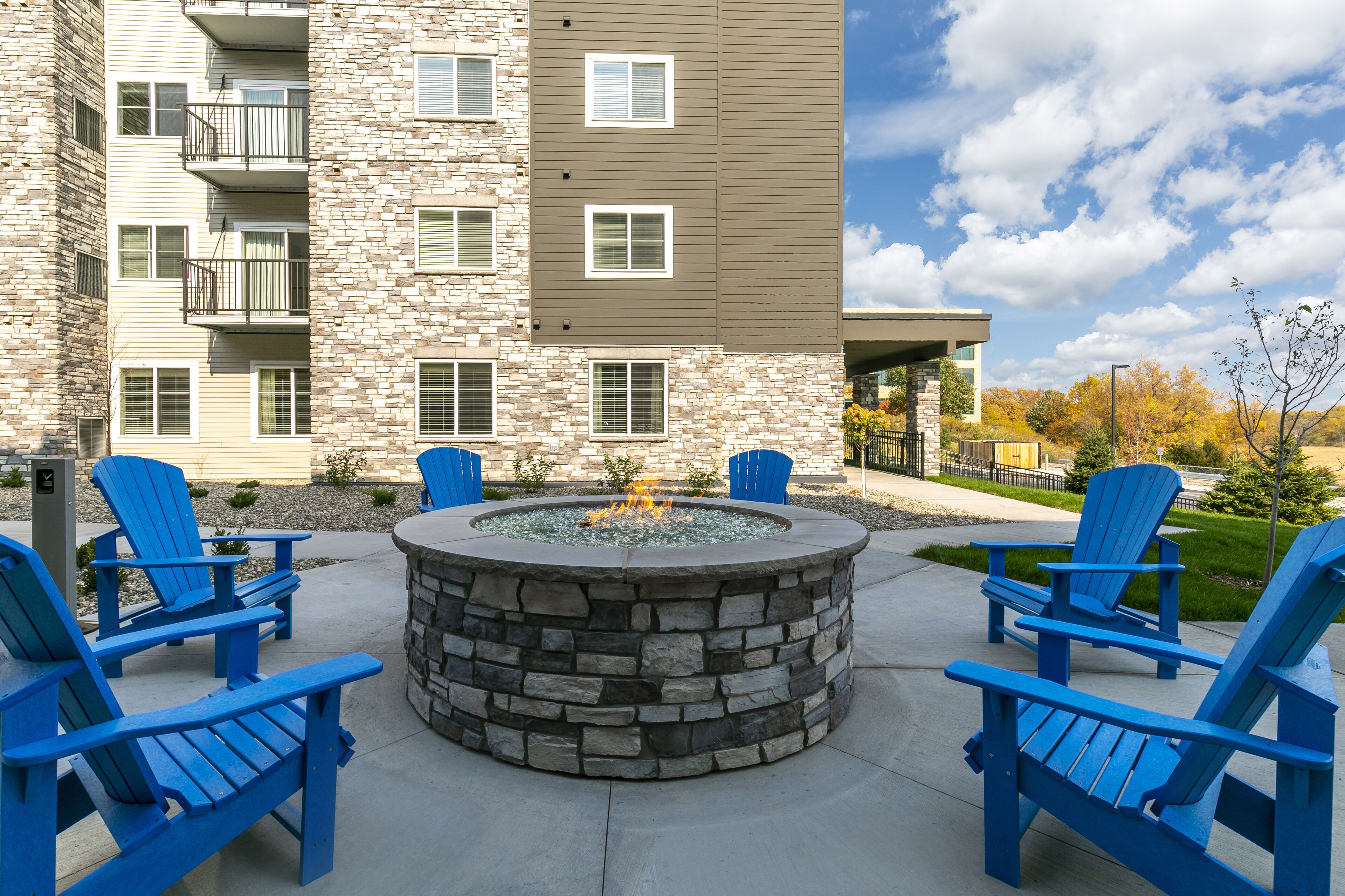 Ein Steinkamin mit vier blauen Adirondack-Stühlen darum, auf einer Betonterrasse vor einem mehrstöckigen Gebäude mit Balkonen.