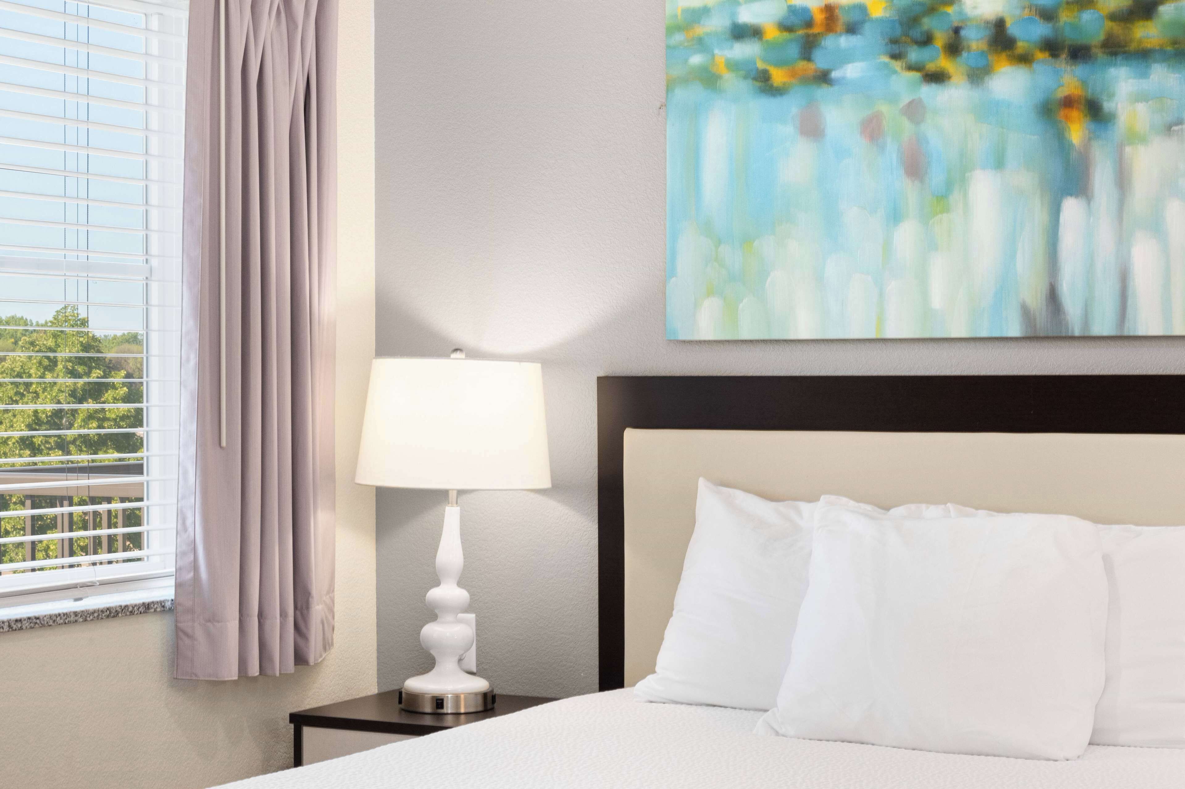 A hotel room with a bed, a nightstand, a lamp, a window with curtains, and a painting on the wall.