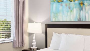 A hotel room with a bed, a nightstand, a lamp, a window with curtains, and a painting on the wall.