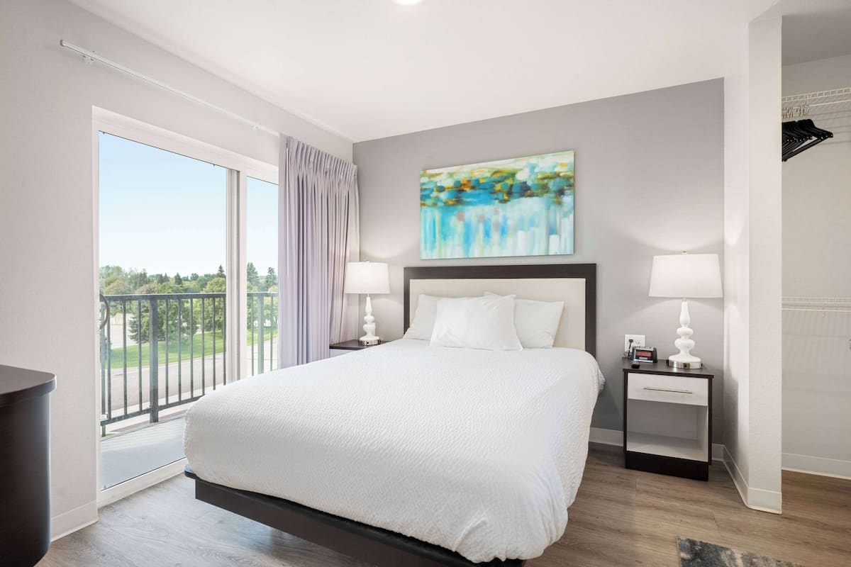 A modern hotel room with a large bed, two bedside tables with lamps, a painting on the wall, and a balcony with a view.