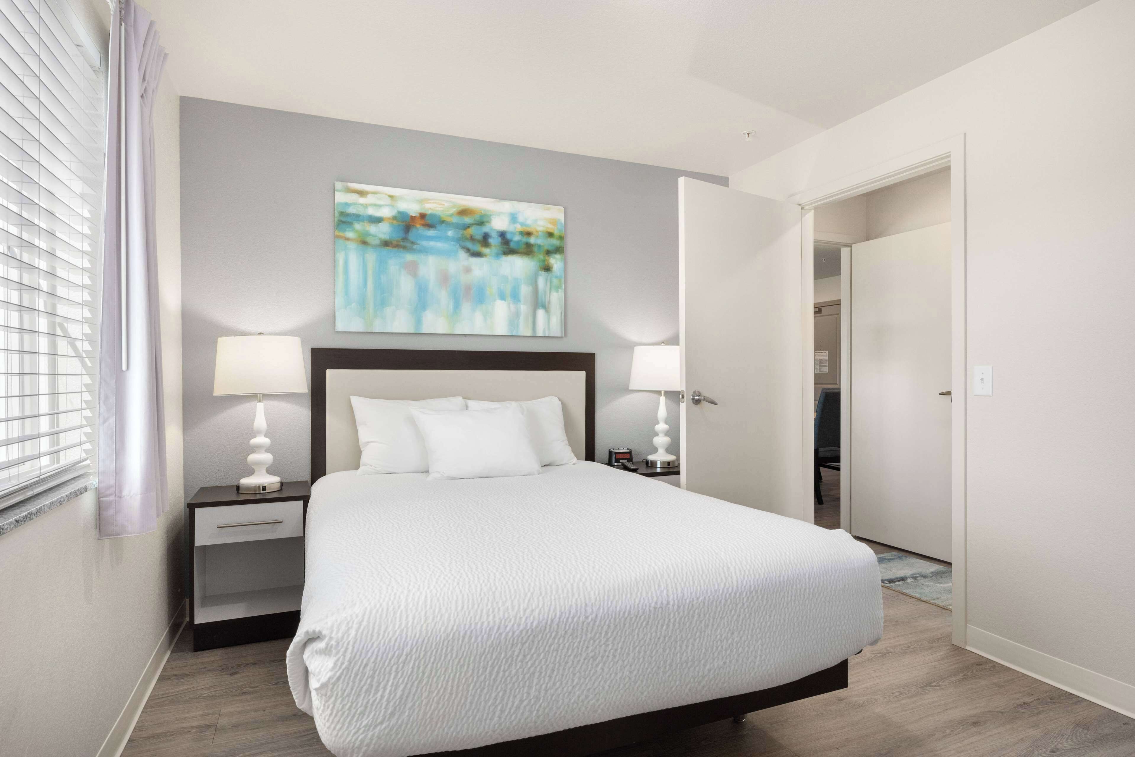 Quarto de hotel moderno com uma cama grande, duas mesas de cabeceira com luminárias, um quadro na parede e uma porta que dá para outro quarto.