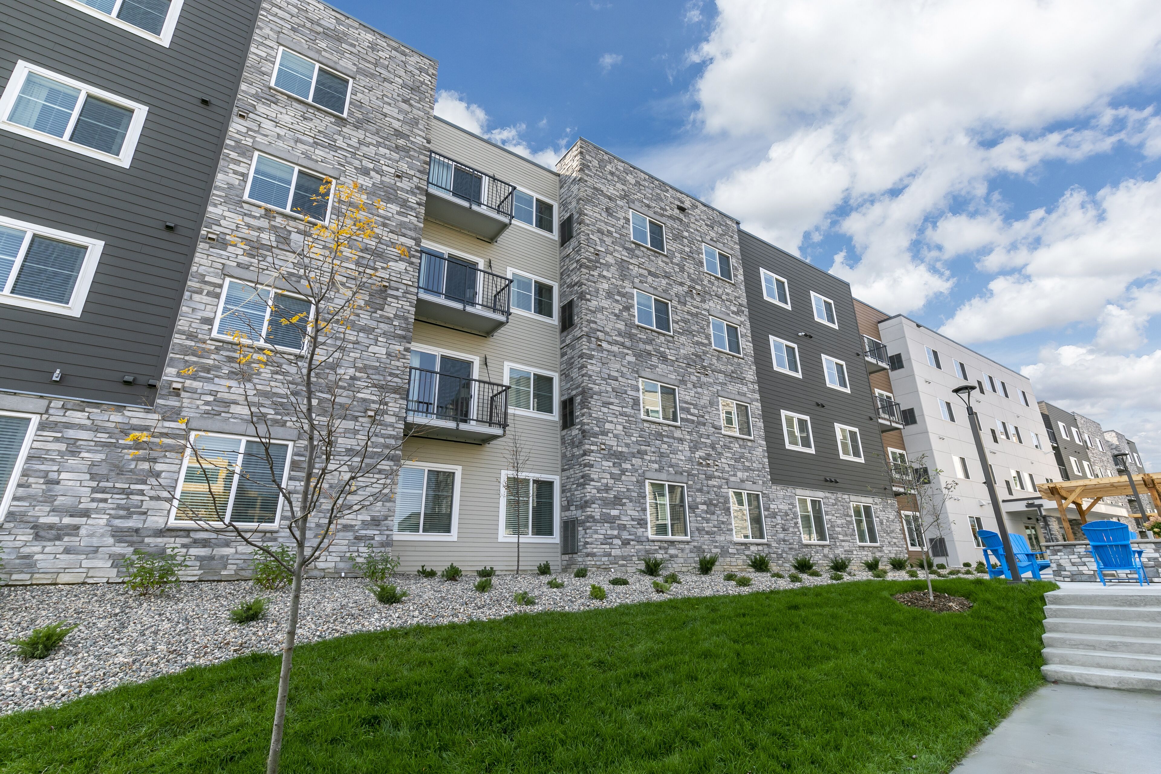 Un edificio residencial de varios pisos con balcones, un patio delantero con césped y una zona de estar exterior azul.