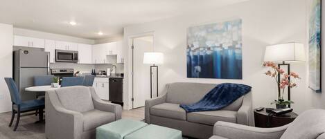 A modern living room with a grey sofa, two armchairs, and a matching ottoman. A dining area with a white table and blue chairs is visible in the background.