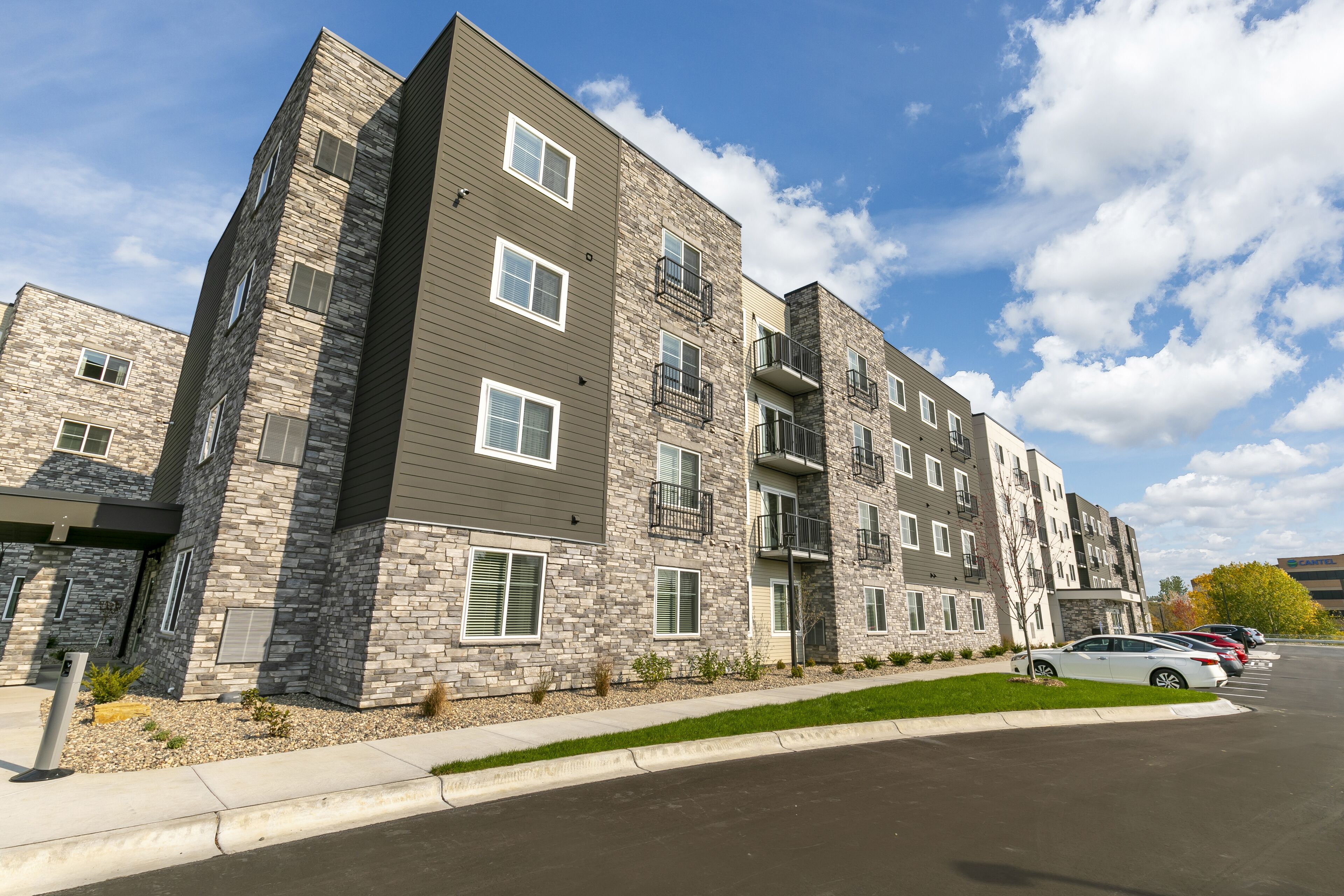 A multi-story apartment building with a stone facade, balconies, and a parking lot with cars.