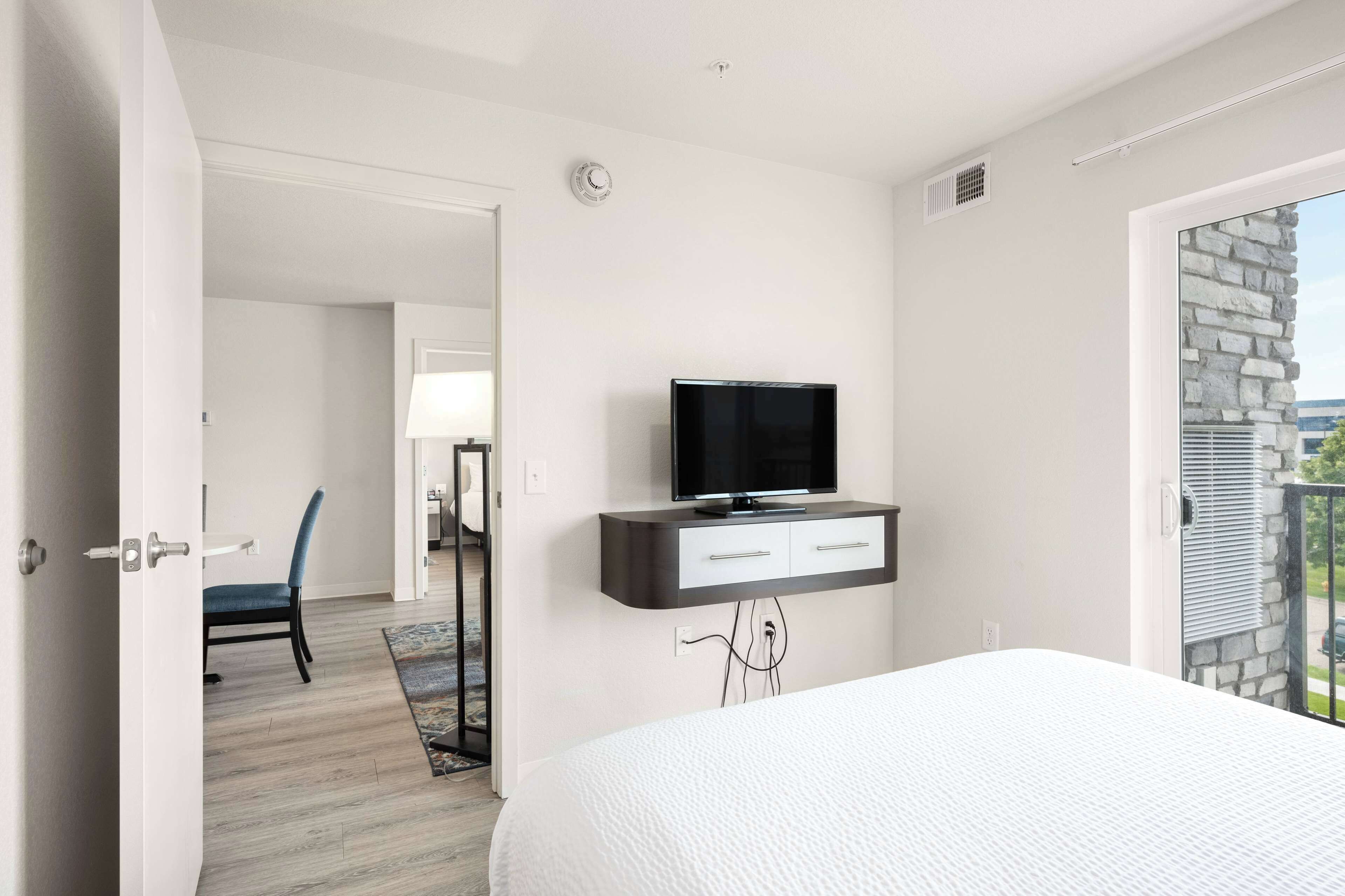A hotel room with a bed, a TV mounted on a wall, a door leading to a balcony, and a chair in the corner.