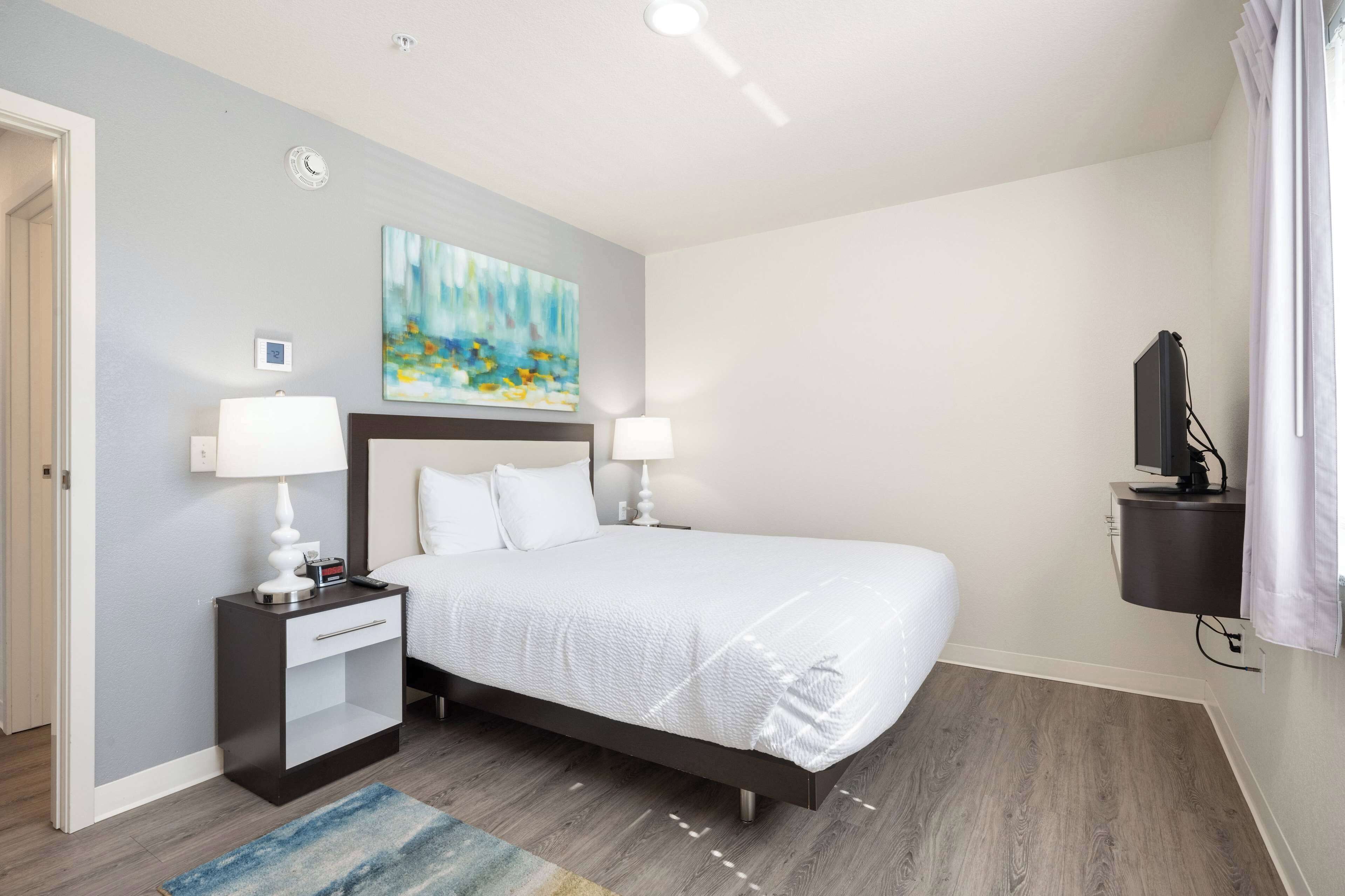 Habitación de hotel con cama, lámparas de noche, un cuadro, televisor y una mesita.