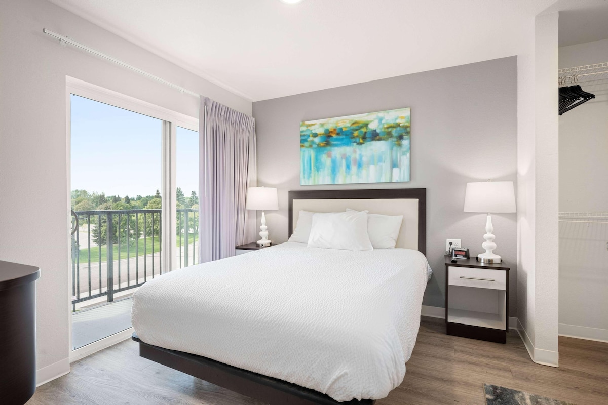 Una habitación de hotel moderna con una cama grande, dos mesitas de noche, un cuadro y un balcón con vistas.