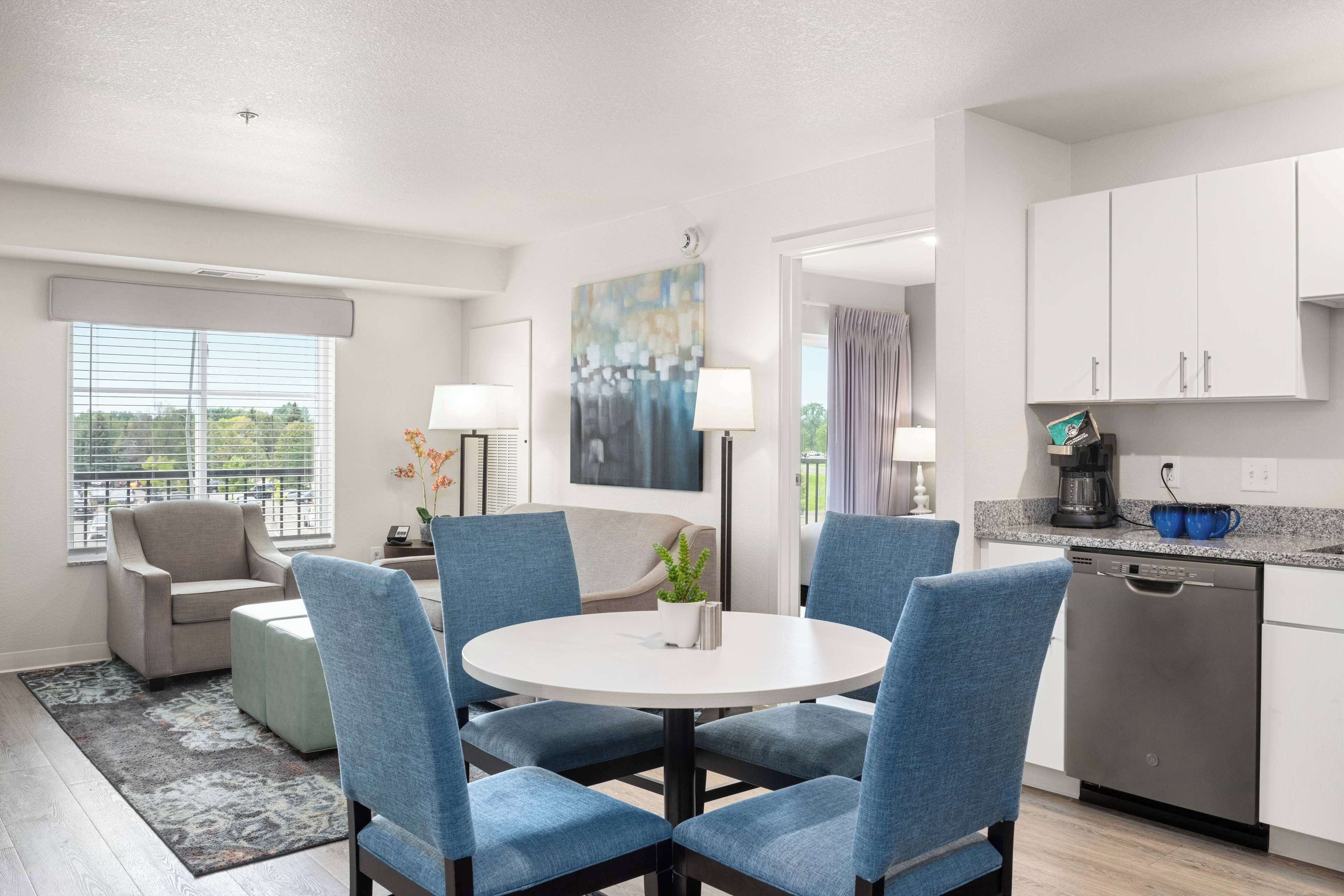 A modern kitchen with white cabinets, a dishwasher, and a coffee maker. There is a round table with blue chairs and a small potted plant.