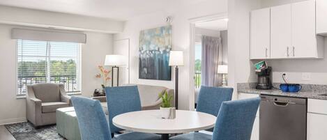 A modern kitchen with white cabinets, a dishwasher, and a coffee maker. There is a round table with blue chairs and a small potted plant.