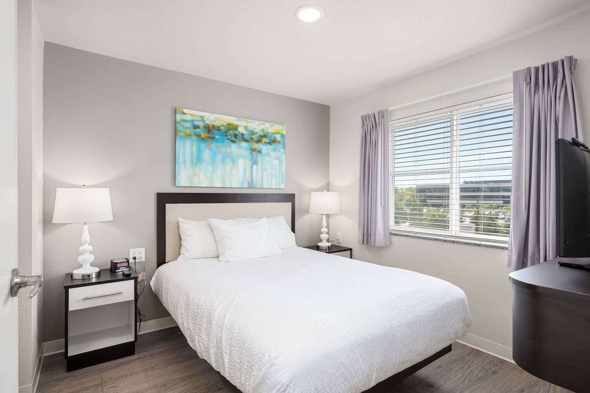 A hotel room with a large bed, two bedside tables with lamps, a painting on the wall, a window with curtains, and a television on a stand.
