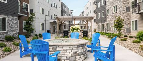 Une cour avec un foyer en pierre, entourée de chaises Adirondack bleues, une pergola et une table à manger avec des chaises.