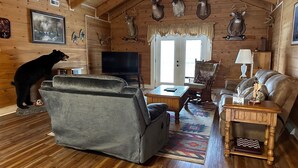 Interior de una cabaña de troncos con un sofá, un sillón, un televisor y cornamentas de ciervo en la pared.