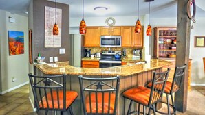 Una cocina con isla central, taburetes de bar, armarios de madera e iluminación colgante.