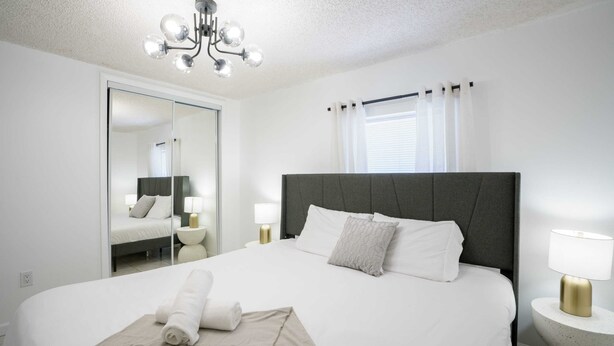 A hotel room with a large bed, a mirror, a chandelier, and a window with curtains. - Modern 3-Bedroom in Heart of Wynwood Art District (Miami)