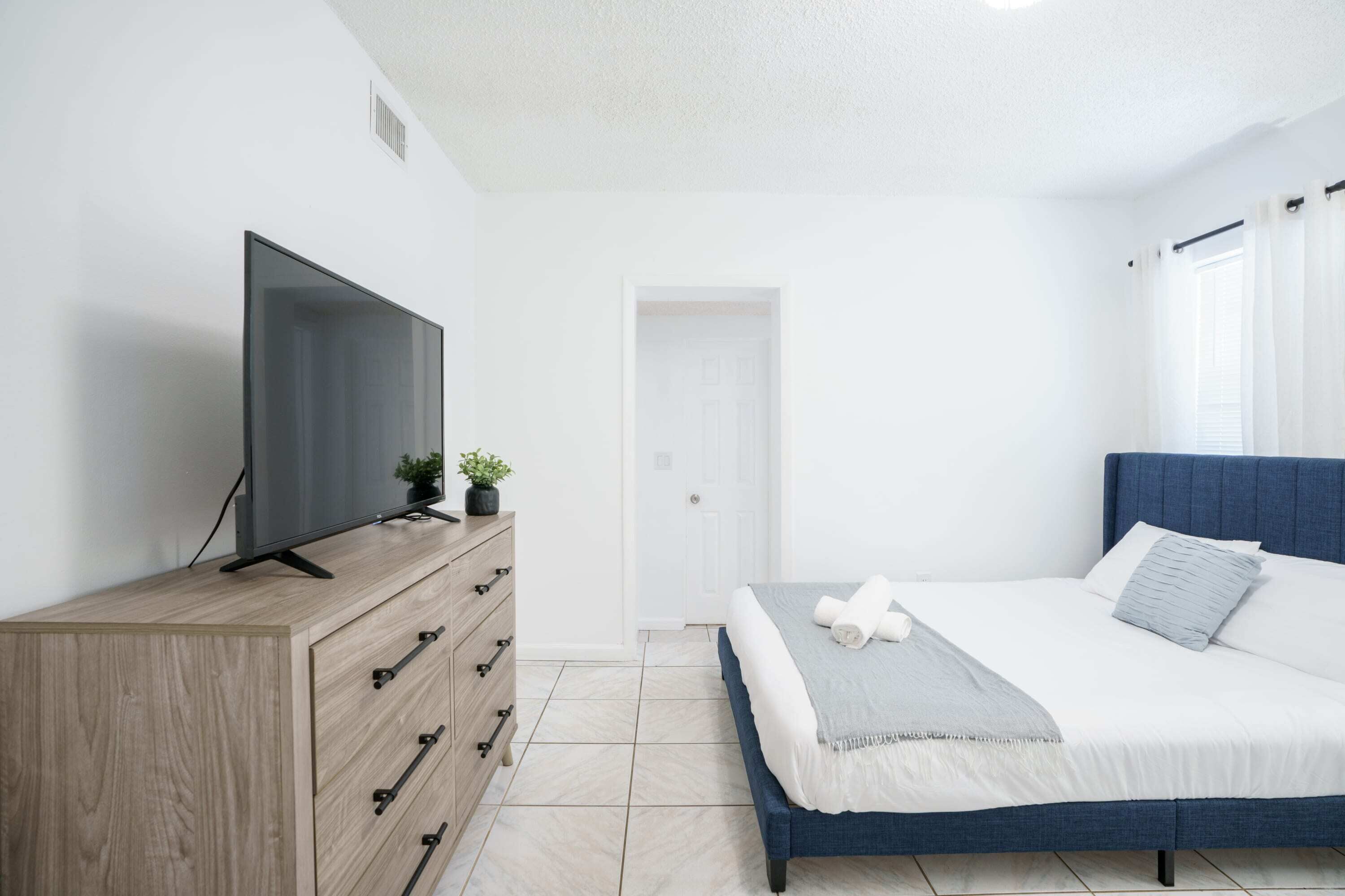 Ein modernes Schlafzimmer mit einem Bett, einem Fernseher auf einer Holzkommode und einer Tür im Hintergrund.