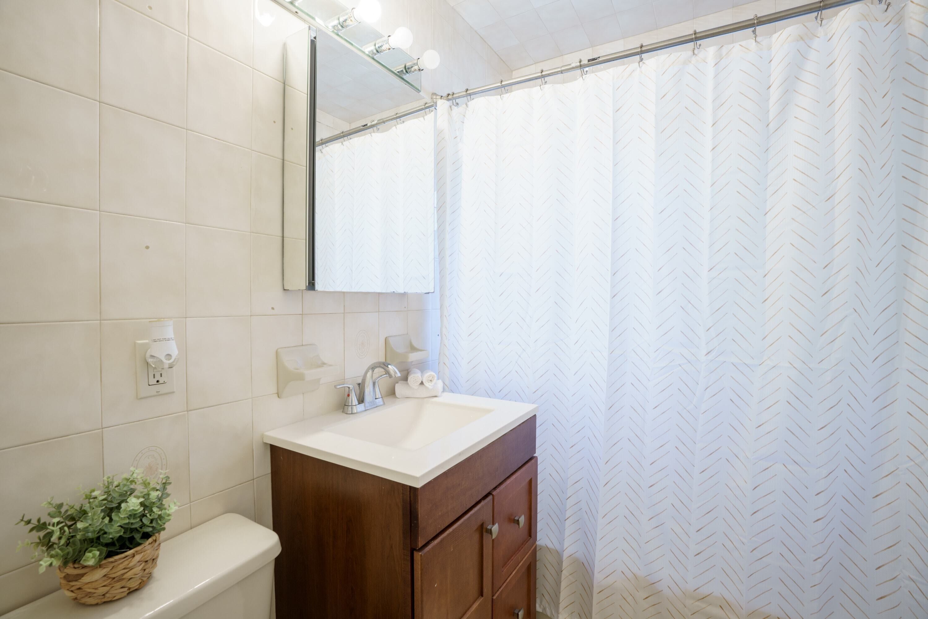 Un baño con lavamanos blanco, tocador de madera, un espejo y cortina de ducha.