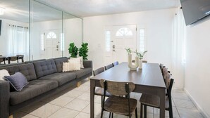 A modern living room with a grey sofa, a wooden dining table, and chairs. There is a large mirror on one wall and a flat-screen TV mounted above the table. - Modern 3-Bedroom in Heart of Wynwood Art District (Miami)