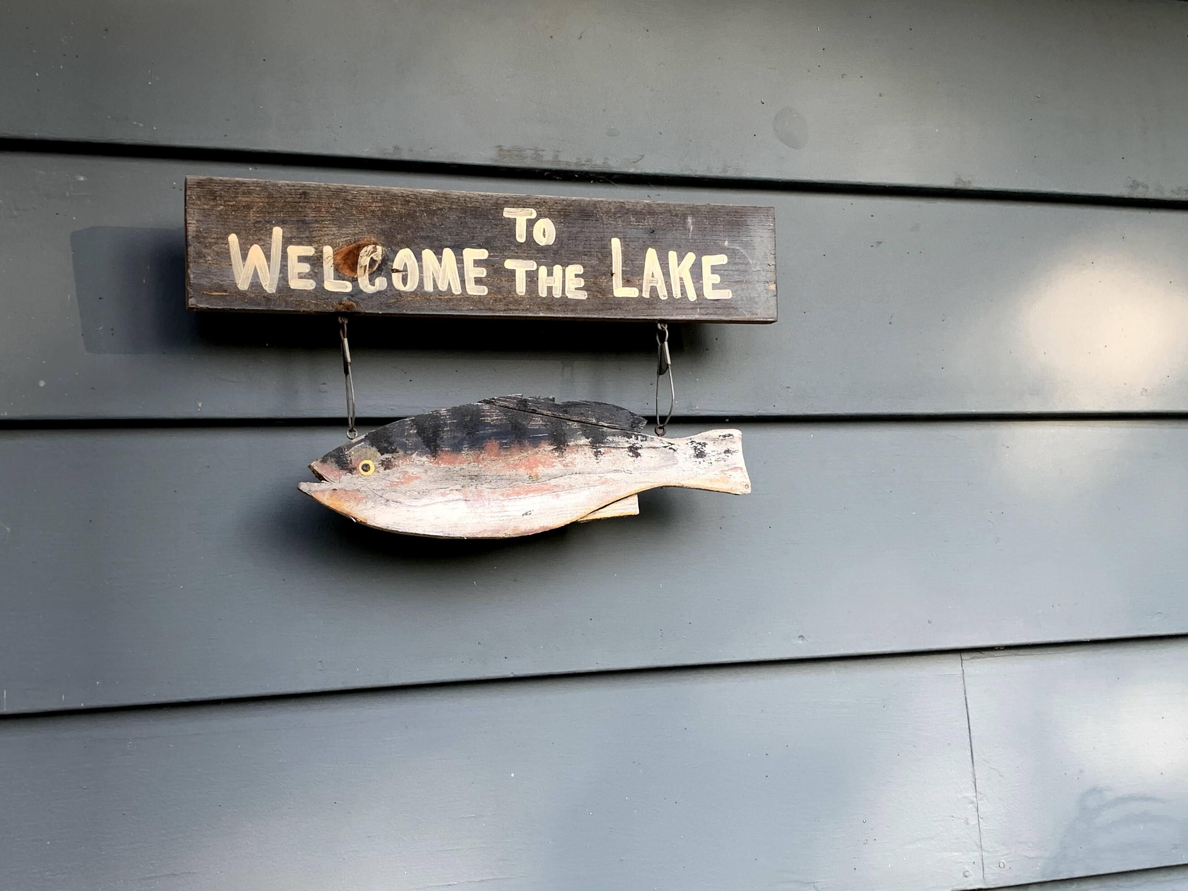 A wooden sign with the text "To Welcome the Lake" hanging on a gray wall.