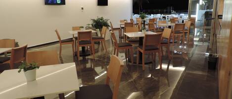 A modern dining area with multiple tables and chairs, a flat-screen TV mounted on the wall, and a potted plant on one table.
