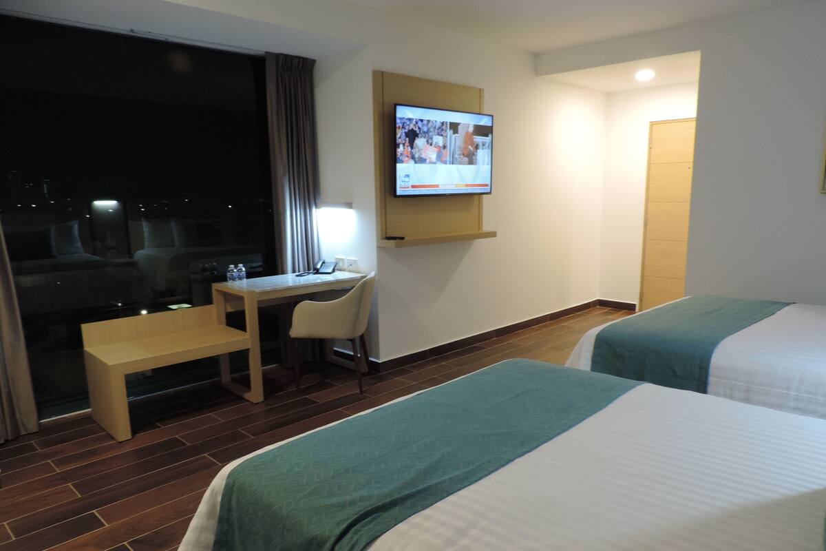 A hotel room with two beds, a desk, a chair, a TV mounted on the wall, and a window with curtains.