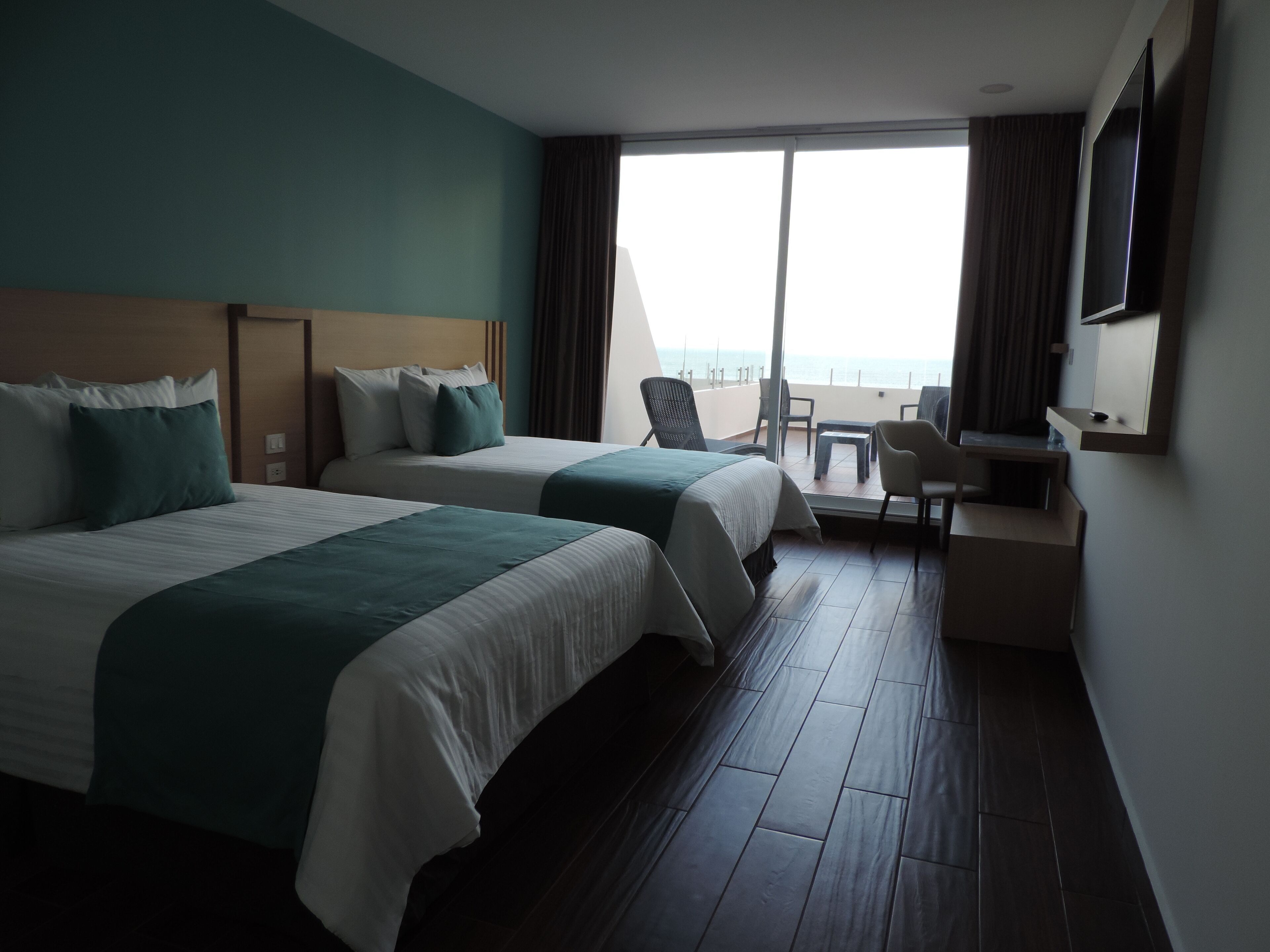 A hotel room with two beds, a desk, a chair, and a large window with a view of the sea.