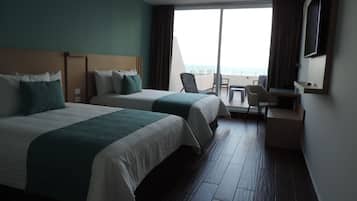 A hotel room with two beds, a desk, a chair, and a large window with a view of the sea.