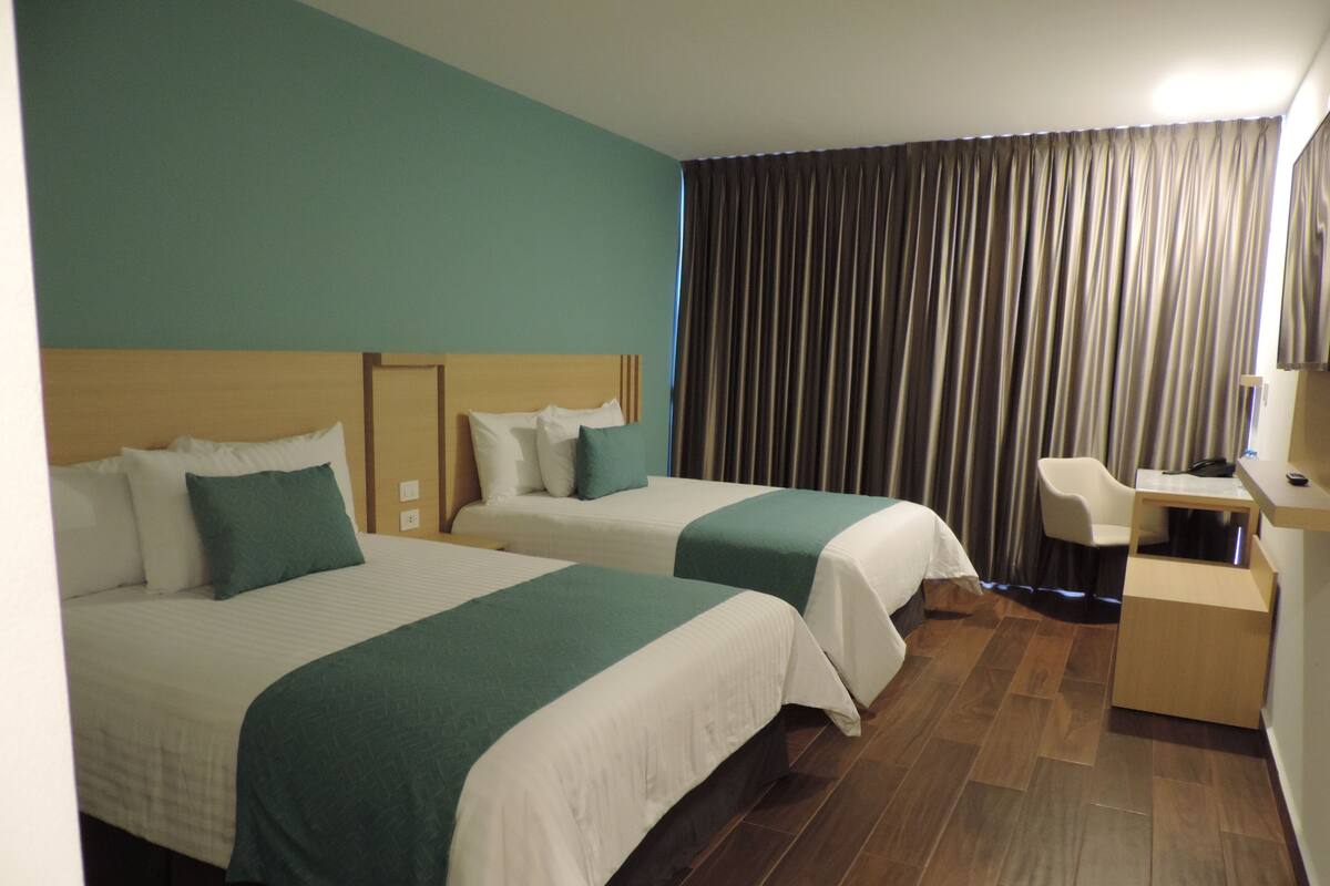 A hotel room with two beds, a desk, a chair, and a large window with curtains.