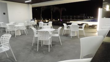 A spacious outdoor area with white plastic chairs and tables arranged for dining, a pool in the background, and a dark night sky.