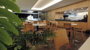 Un comedor moderno con mesas y sillas de madera, plantas en macetas y una cocina al fondo.