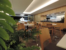 Un comedor moderno con mesas y sillas de madera, plantas en macetas y una cocina al fondo.