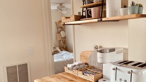 A kitchen with wooden shelves, a coffee maker, a toaster, and a basket of bread. - Comfy Cottage With Writer Cabin and Book Nook. Pet-Friendly Fully Fenced Yard (Joshua Tree)