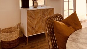 A room with a herringbone-patterned cabinet, a wicker chair, a woven basket, and a view of a desert landscape through a window. - Comfy Cottage With Writer Cabin and Book Nook. Pet-Friendly Fully Fenced Yard (Joshua Tree)