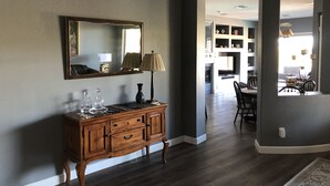 A room with a wooden console table, a mirror, and a lamp. There is a doorway leading to another room with a dining table and chairs.