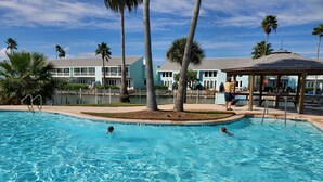 Una piscina azul cristalina con dos personas nadando, rodeada de palmeras y un pabellón.