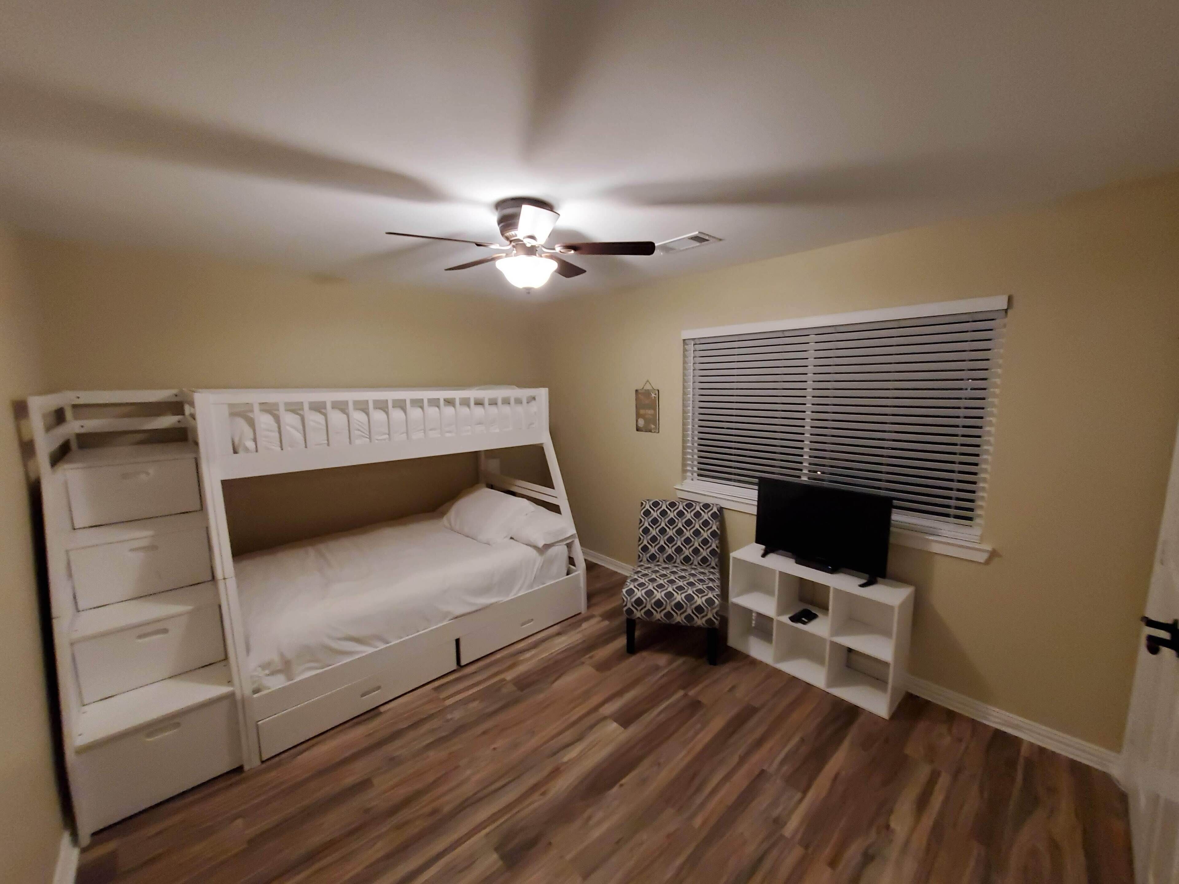 Un dormitorio con literas, ventilador de techo, televisor sobre un soporte, ventana con persianas y una silla estampada.