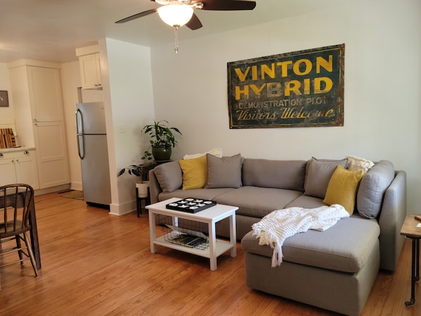 A living room with a grey sofa, wooden floor, white walls, a ceiling fan, and a Vinton Hybrid sign.