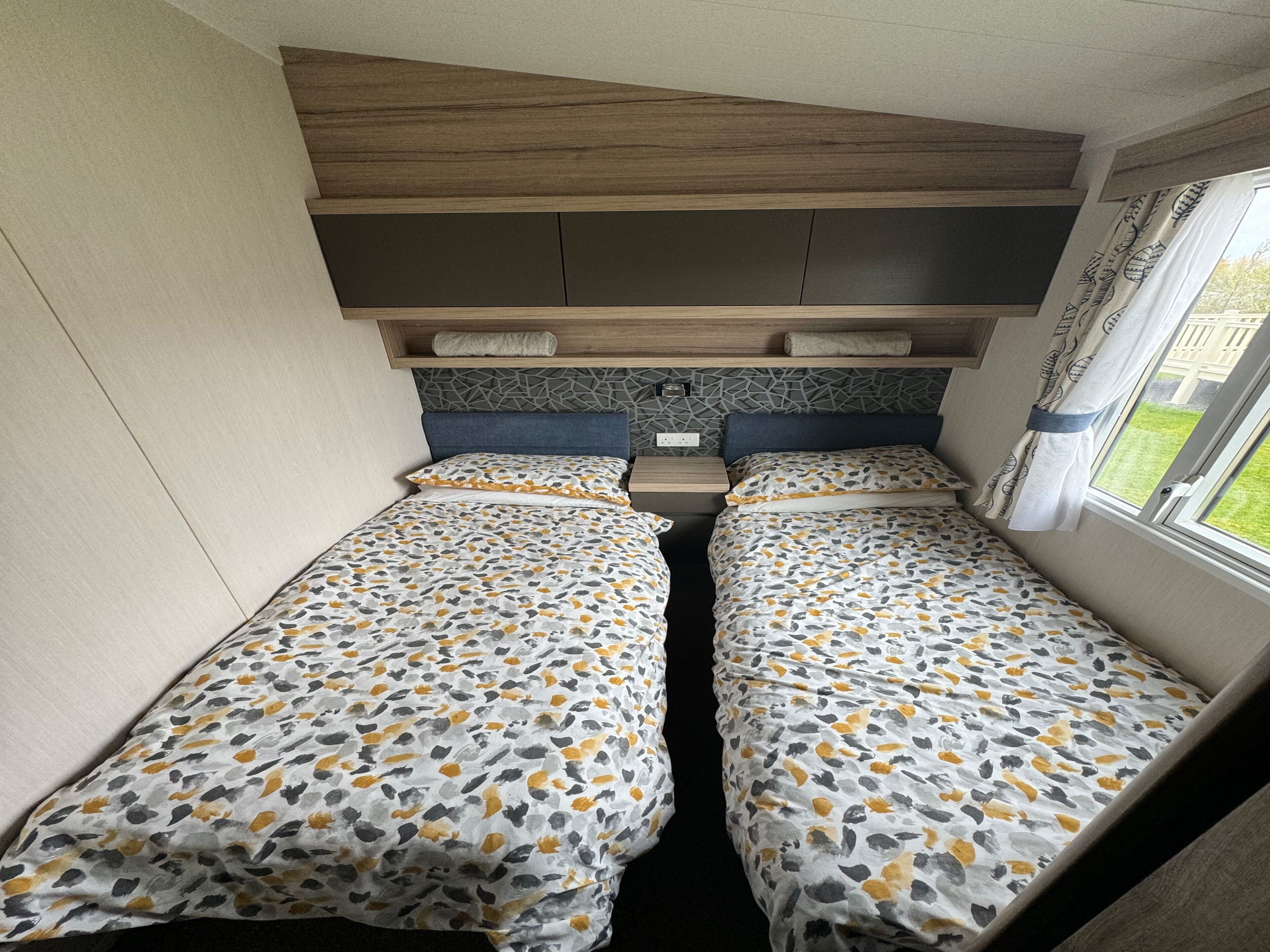 A compact RV bedroom with two beds, a built-in headboard, and a window with curtains.