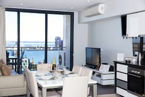 A modern living room with a dining table set for four, a sofa, a TV, and a kitchen area with a stove and oven.