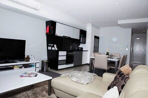 A modern living room with a flat-screen TV, a sofa, a coffee table, and a kitchen area with cabinets and a microwave.