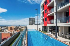A rooftop swimming pool with a city view, a red building, and a tall structure in the background.