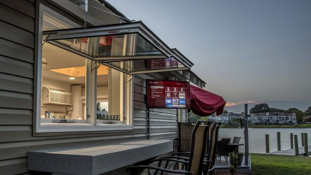 A deck with outdoor seating, a TV screen displaying channel information, and a view of the water. - Riverside Tiny Home Retreat  (Hampton)