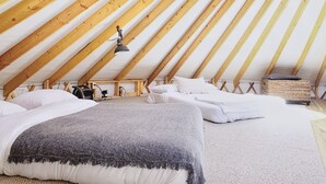 Un dormitorio acogedor en el ático, con techo inclinado, vigas de madera, una cama con sábanas blancas, una manta gris y una mesita con una lámpara.